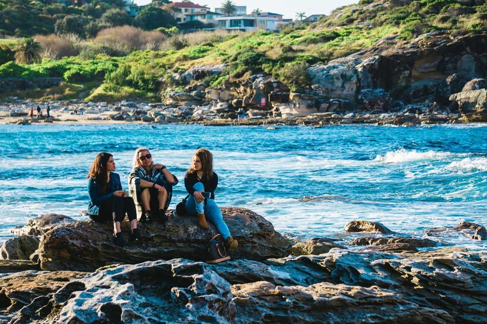 tres chicas sentadas en una roca en el mar y detrás la vegetación